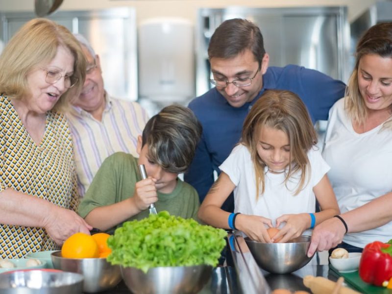 Family of various ages gathers in kitchen to cook meal together, showcasing culinary togetherness.