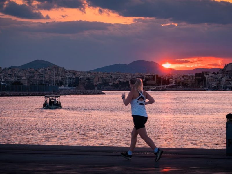 A woman jogs along a waterfront at sunset with a boat in the background.